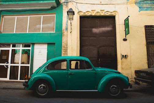 A classic green Volkswagen Beetle is parked along a street in front of a building with green and beige walls. The building features a large dark brown door and modern window design. An old street lamp hangs above the car, and there are other parked vehicles partially visible.