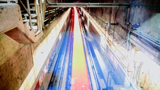 A tunnel with industrial design featuring concrete walls and overhead pipes. A bright red and blue path leads through the center, flanked by metal railings and illuminated by artificial lighting.