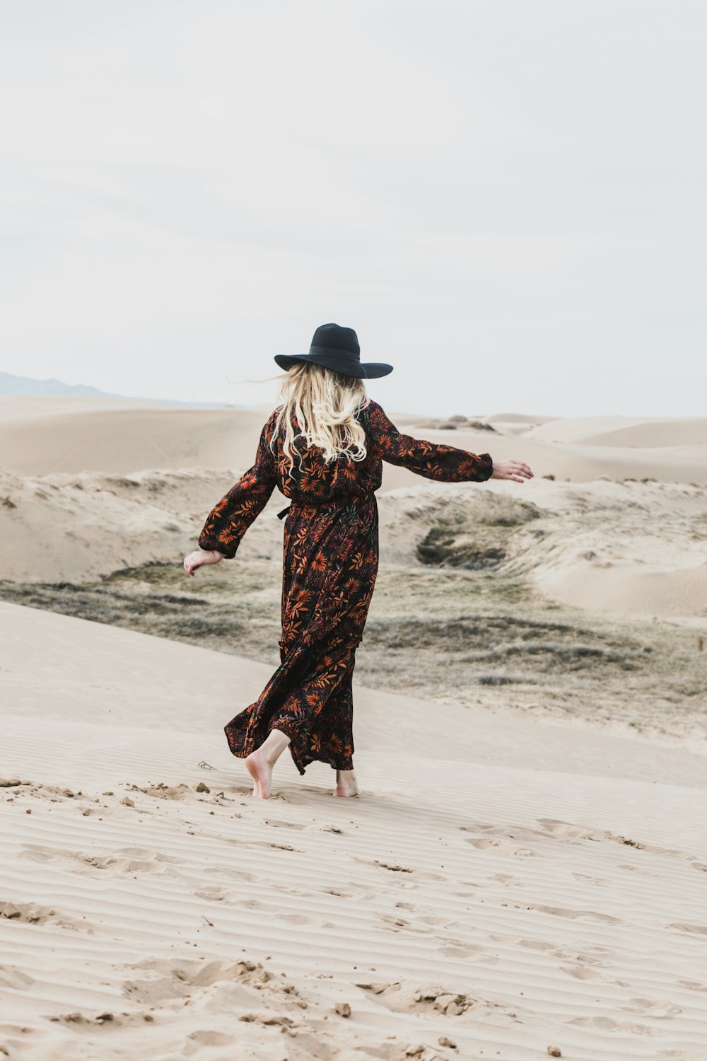 Foto einer Person in einem braunen und orangefarbenen Maxikleid mit Blumenmuster, die barfuß durch verlassenes Land geht