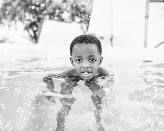 A young child is partially submerged in water, looking directly at the camera. The surface of the water has sparkling reflections with visible bubbles. The background is blurred, focusing attention on the child.