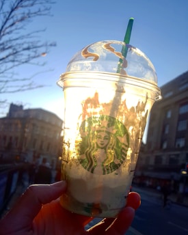 A partially consumed Starbucks beverage is held up against a backdrop of a city street during sunset. The sunlight shines through the plastic cup, casting a warm glow. The iconic green logo is prominently visible on the cup. The background displays urban buildings and a clear blue sky.