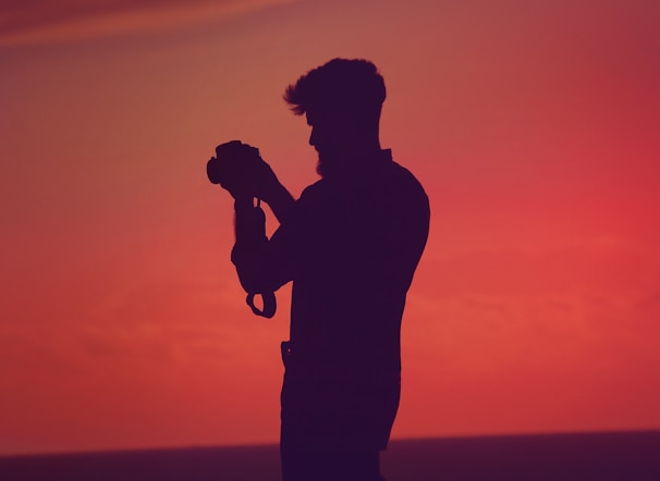 A silhouette of a person holding a camera against a vibrant sunset background. The scene is dominated by deep reds and oranges, creating a striking and dramatic backdrop. The figure appears to be focused on capturing the moment, highlighting a passion for photography.