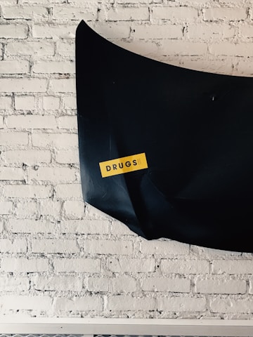 A black object is mounted on a white brick wall with a yellow sticker displaying the word 'DRUGS' in bold letters.