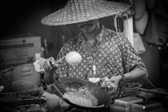 A person wearing a large woven hat and a patterned shirt stands at a food stall, holding a ladle over a wok-like pan. Various cooking ingredients and utensils are visible in the foreground, with baskets and stacked trays around the area. The scene suggests a street market or food festival setting.