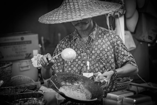 A person wearing a large woven hat and a patterned shirt stands at a food stall, holding a ladle over a wok-like pan. Various cooking ingredients and utensils are visible in the foreground, with baskets and stacked trays around the area. The scene suggests a street market or food festival setting.