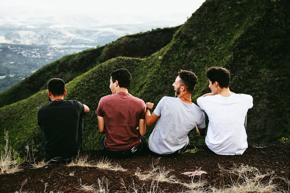 A group of four laughing men sits on an edge overlooking a green valley