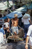 A bustling outdoor market scene depicting a shirtless man in a straw hat carrying a woven basket with corn. He is surrounded by other people, including a woman holding a child, engaged in conversation or transaction. The setting appears to be rustic, with metal-roofed buildings and trees providing shade.