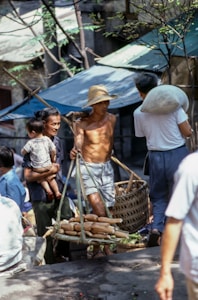 A bustling outdoor market scene depicting a shirtless man in a straw hat carrying a woven basket with corn. He is surrounded by other people, including a woman holding a child, engaged in conversation or transaction. The setting appears to be rustic, with metal-roofed buildings and trees providing shade.