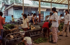 A busy outdoor market scene with people interacting and engaging in trade. Individuals are gathered around carts filled with fresh produce, such as leafy greens and melons. Some people are standing while others are seated on structures around the area, giving a sense of casual social interaction.