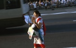 A person walking on a street holding a map and several brochure-like items, surrounded by fast-moving traffic. The background consists of a busy road and spectators sitting along an elevated walkway.