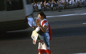 A person walking on a street holding a map and several brochure-like items, surrounded by fast-moving traffic. The background consists of a busy road and spectators sitting along an elevated walkway.