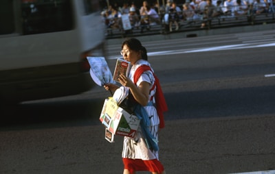 A person walking on a street holding a map and several brochure-like items, surrounded by fast-moving traffic. The background consists of a busy road and spectators sitting along an elevated walkway.