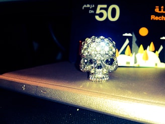 A skull-shaped ornament adorned with sparkling rhinestones rests on a textured surface. Behind the skull, there is a card featuring the text 'Dhs 50' and an abstract graphic of geometric shapes and trees.