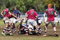 A group of rugby players in action on a grass field, wearing jerseys with red, white, and blue colors. The players appear to be engaged in a tackle, surrounded by others ready to support or oppose the movement. The ground is slightly muddy, indicating recent play or rain. Trees and parked cars are visible in the background.