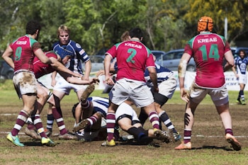 A group of rugby players in action on a grass field, wearing jerseys with red, white, and blue colors. The players appear to be engaged in a tackle, surrounded by others ready to support or oppose the movement. The ground is slightly muddy, indicating recent play or rain. Trees and parked cars are visible in the background.