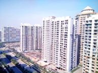 High-rise residential buildings dominate the landscape with multiple apartment units visible. The street below is flanked by green trees and a few vehicles are parked along the roadside. The sky above is clear and blue, giving an impression of a sunny day.