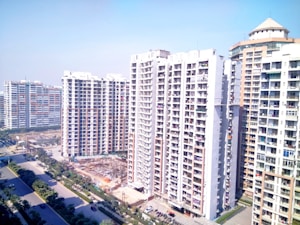 High-rise residential buildings dominate the landscape with multiple apartment units visible. The street below is flanked by green trees and a few vehicles are parked along the roadside. The sky above is clear and blue, giving an impression of a sunny day.