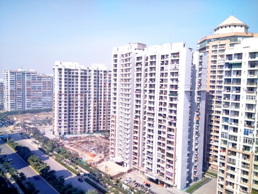 High-rise residential buildings dominate the landscape with multiple apartment units visible. The street below is flanked by green trees and a few vehicles are parked along the roadside. The sky above is clear and blue, giving an impression of a sunny day.