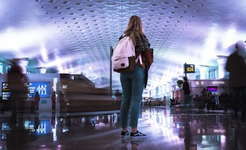 A person stands in an airport terminal with a backpack, surrounded by blurred figures representing movement. The ceiling is illuminated with geometric patterns, and various signs and displays are visible, including flight information and advertisements.