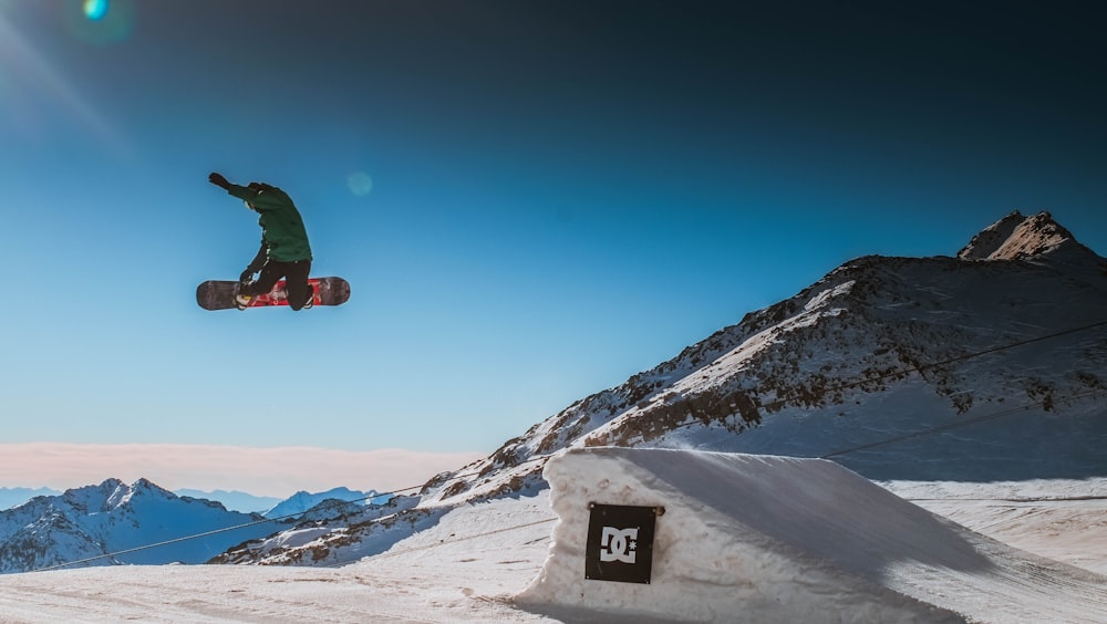 person wearing green jacket jumping on white snow ramp during daytime