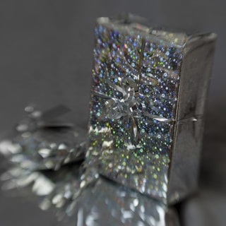 A shiny, holographic gift box with a star pattern, topped with a reflective silver ribbon, stands in front of a similar, partially visible box, against a dark, muted background.