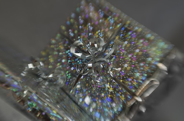 A close-up of a shiny gift box adorned with a holographic star-patterned wrapping paper. The box is topped with a reflective silver bow that catches light, creating a sparkling effect. The image has a blurred, zoom-effect background, emphasizing the decoration.