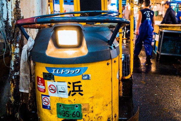 A yellow industrial vehicle is parked in an indoor market setting, with multiple stickers and a license plate attached. Nearby, a person wearing blue overalls walks away, highlighting a busy working environment.