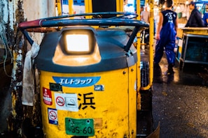 A yellow industrial vehicle is parked in an indoor market setting, with multiple stickers and a license plate attached. Nearby, a person wearing blue overalls walks away, highlighting a busy working environment.