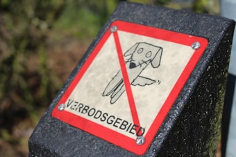 A rectangular warning sign with a red border and a diagonal red line across a drawing of a dog. The word 'VERBODSGEBIED' is printed at the bottom. The sign is attached to a textured black post.