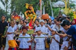 A vibrant cultural parade features a group of children dressed in traditional attire, carrying a colorful effigy. The children, along with adults, are engaged and smiling. Various traditional garments, including headscarves and sarongs, are visible. The crowd in the background shows onlookers, some holding up phones or cameras to capture the moment. Trees and festival decorations line the street.