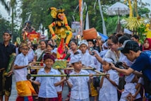 A vibrant cultural parade features a group of children dressed in traditional attire, carrying a colorful effigy. The children, along with adults, are engaged and smiling. Various traditional garments, including headscarves and sarongs, are visible. The crowd in the background shows onlookers, some holding up phones or cameras to capture the moment. Trees and festival decorations line the street.