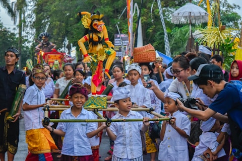 A vibrant cultural parade features a group of children dressed in traditional attire, carrying a colorful effigy. The children, along with adults, are engaged and smiling. Various traditional garments, including headscarves and sarongs, are visible. The crowd in the background shows onlookers, some holding up phones or cameras to capture the moment. Trees and festival decorations line the street.