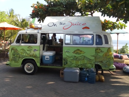 A vintage van painted in green with artistic designs is parked under a lush tree by the seaside. The van has a sign on top that reads 'On the Juice' and appears to be a smoothie or juice bar with supplies like a cooler and water bottles placed outside. The surroundings include tropical plants and a few seating arrangements on the patio.
