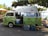 A vintage van painted in green with artistic designs is parked under a lush tree by the seaside. The van has a sign on top that reads 'On the Juice' and appears to be a smoothie or juice bar with supplies like a cooler and water bottles placed outside. The surroundings include tropical plants and a few seating arrangements on the patio.