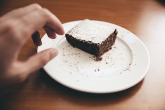 A piece of chocolate brownie dusted with powdered sugar rests on a white plate. A hand is reaching towards the dessert, suggesting someone is about to take it. Crumbs are scattered around, and the plate is set on a wooden surface.