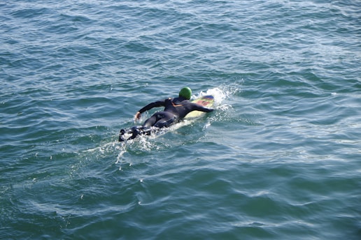 A person wearing a wetsuit and a green swim cap is paddling on a surfboard across the open water. The scene suggests a setting for surfing or ocean sports.