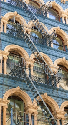 A building facade featuring ornate architectural detailing with arched windows and decorative columns. The structure is adorned with a series of metal fire escapes crossing diagonally in front of the windows. The color scheme includes light blue walls and beige accents around the arches and columns.