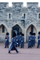 A soldier in ceremonial uniform marches in front of a group of similarly dressed soldiers standing in formation. The soldiers are wearing tall black hats and long grey coats. Behind them is a historic stone building with arched windows.