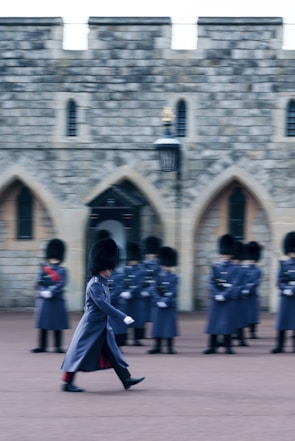 A soldier in ceremonial uniform marches in front of a group of similarly dressed soldiers standing in formation. The soldiers are wearing tall black hats and long grey coats. Behind them is a historic stone building with arched windows.