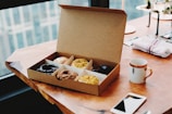 A warm morning scene with a box of donuts beside a steaming cup of coffee on a wooden table.