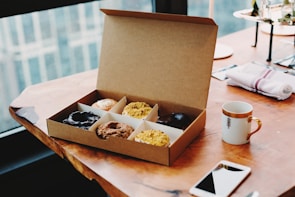 A warm morning scene with a box of donuts beside a steaming cup of coffee on a wooden table.