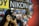 A person with long black hair is taking a photograph using a Nikon camera with a flash. The background features a collage of vibrant photographs and a prominent sign with the word 'NIKON'.