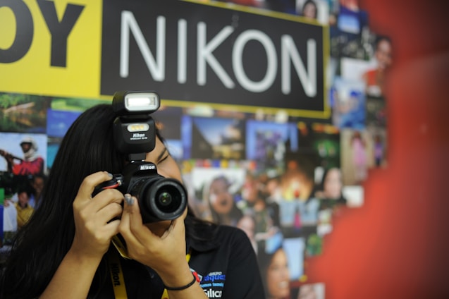A person with long black hair is taking a photograph using a Nikon camera with a flash. The background features a collage of vibrant photographs and a prominent sign with the word 'NIKON'.