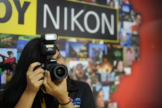 A person with long black hair is taking a photograph using a Nikon camera with a flash. The background features a collage of vibrant photographs and a prominent sign with the word 'NIKON'.