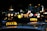 Brightly lit taxi signs are visible on top of vehicles in an urban setting during nighttime. The background features blurred city lights, suggesting a bustling street.