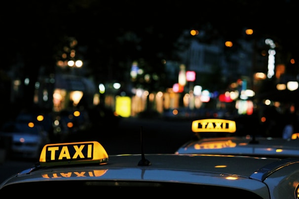 Brightly lit taxi signs are visible on top of vehicles in an urban setting during nighttime. The background features blurred city lights, suggesting a bustling street.
