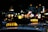 Brightly lit taxi signs are visible on top of vehicles in an urban setting during nighttime. The background features blurred city lights, suggesting a bustling street.