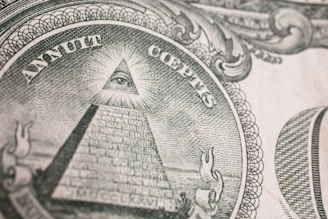 An intricate engraving of the Eye of Providence, commonly known as an all-seeing eye, is depicted within a triangular shape atop an unfinished pyramid. Surrounding the pyramid, there are Latin inscriptions and ornate scrollwork designs, suggesting symbolic and historical significance.