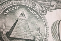 An intricate engraving of the Eye of Providence, commonly known as an all-seeing eye, is depicted within a triangular shape atop an unfinished pyramid. Surrounding the pyramid, there are Latin inscriptions and ornate scrollwork designs, suggesting symbolic and historical significance.