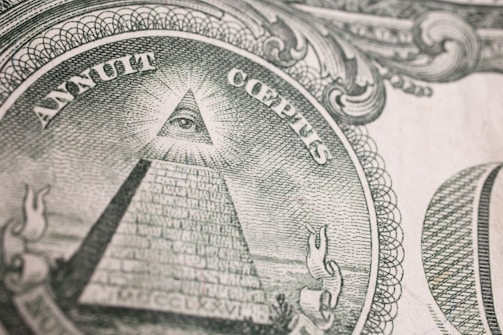An intricate engraving of the Eye of Providence, commonly known as an all-seeing eye, is depicted within a triangular shape atop an unfinished pyramid. Surrounding the pyramid, there are Latin inscriptions and ornate scrollwork designs, suggesting symbolic and historical significance.